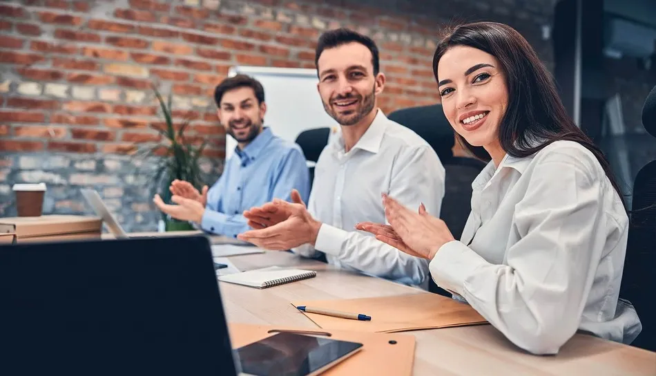 Tres colegas sentados en una mesa, sonriendo y aplaudiendo durante una reunión de negocios, con una laptop en primer plano.